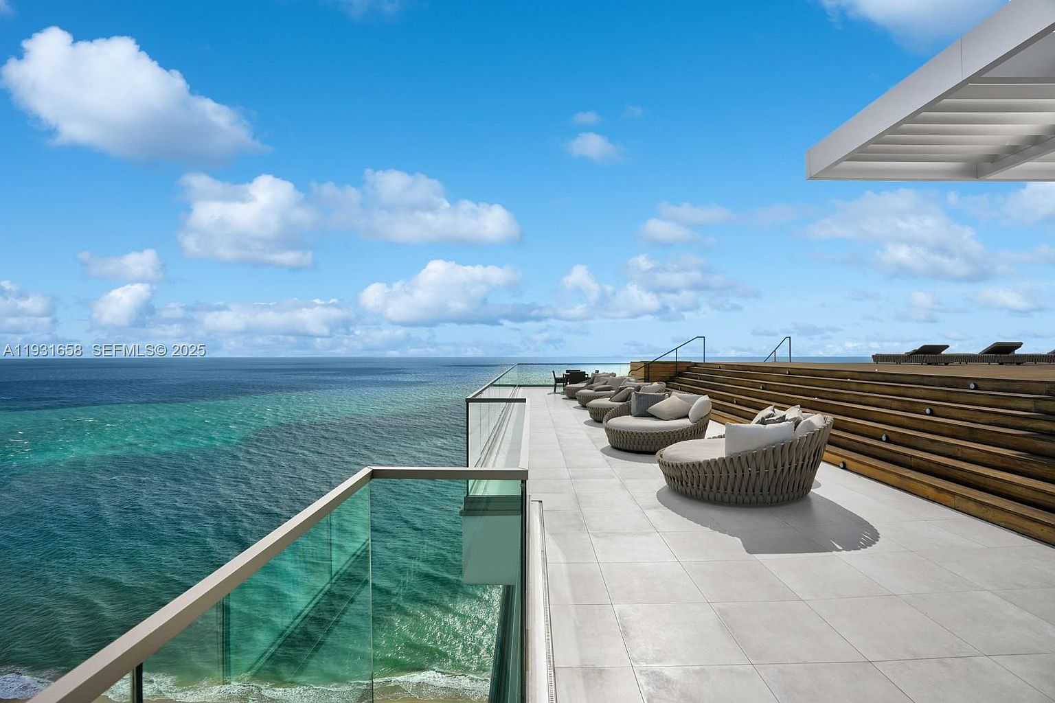 This image showcases a luxurious balcony or deck area with stunning ocean views. The space is furnished with stylish, round wicker seating arrangements, complemented by a modern glass railing. A tiered wooden deck with lounge chairs adds another level of relaxation, creating an inviting outdoor living space perfect for enjoying the coastal scenery.