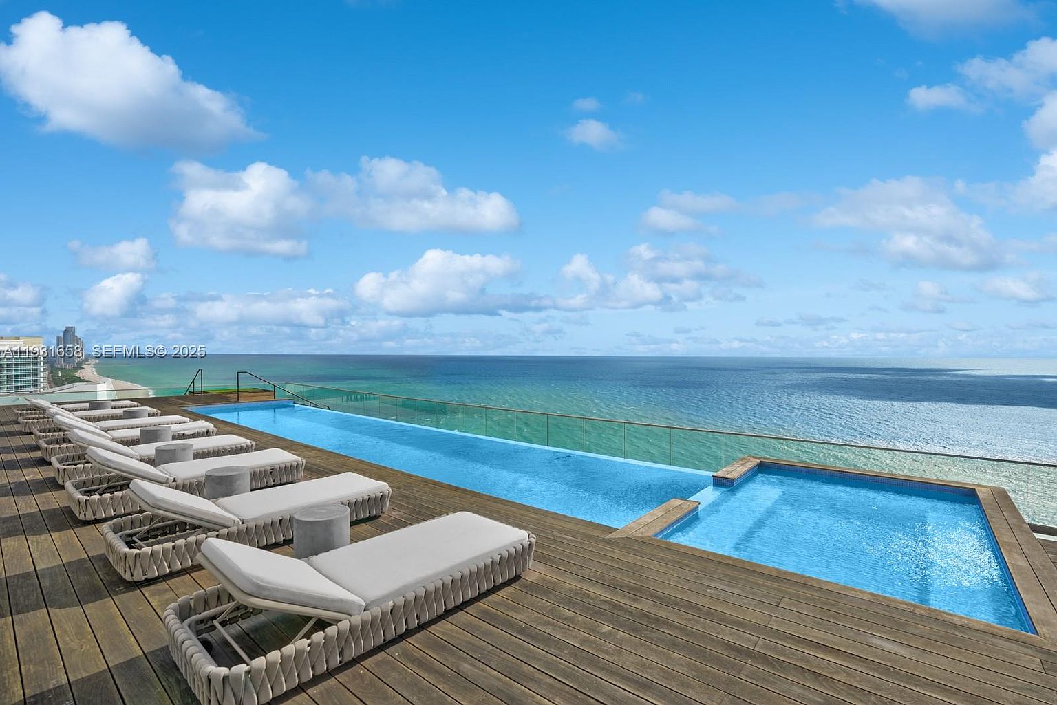 This image showcases a luxurious rooftop pool and spa area with stunning ocean views. The infinity pool blends seamlessly with the horizon, creating a sense of tranquility and exclusivity. Lounge chairs are neatly arranged on the wooden deck, inviting relaxation and enjoyment of the breathtaking scenery.
