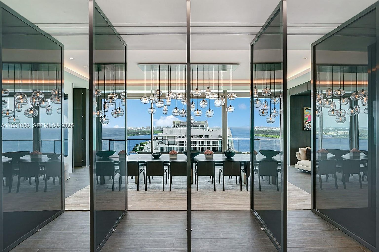 This image showcases a luxurious dining room with a stunning view of the ocean and cityscape. A long, dark wood dining table is surrounded by modern chairs, and a unique chandelier with multiple glass orbs hangs above. The room is framed by dark-framed glass doors, adding to the contemporary and elegant atmosphere.