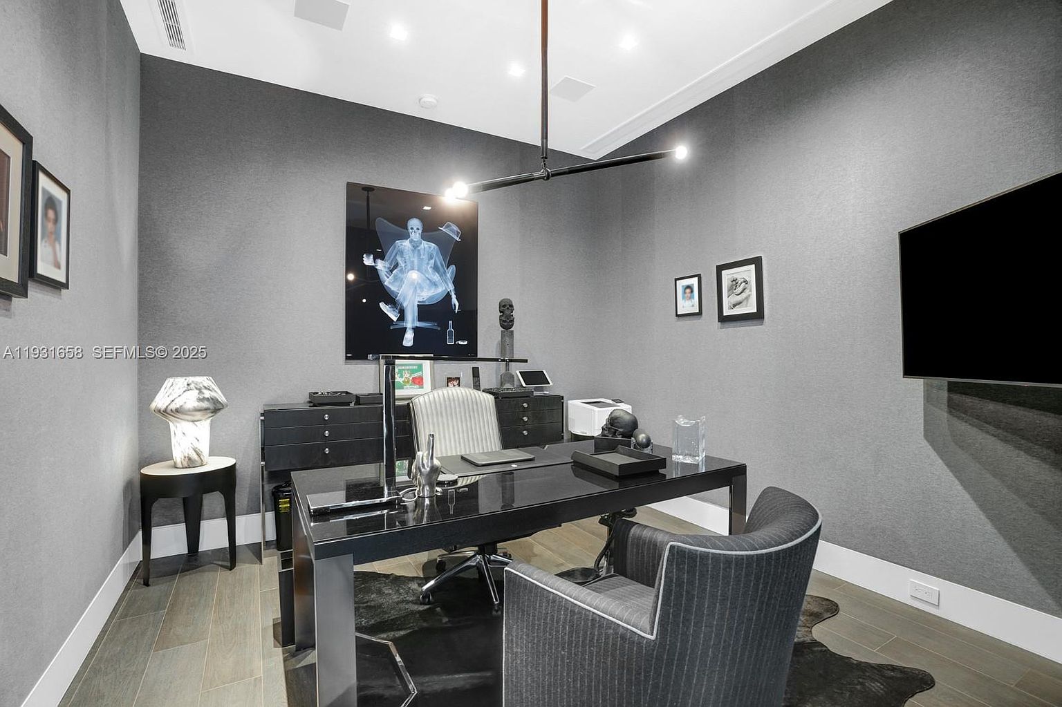 This is a modern office space featuring a black desk, a gray striped chair, and a black and white cowhide rug. The walls are gray, and there is a large piece of art hanging above the desk. The room is well-lit with a modern chandelier and a table lamp, creating a sophisticated and professional atmosphere.