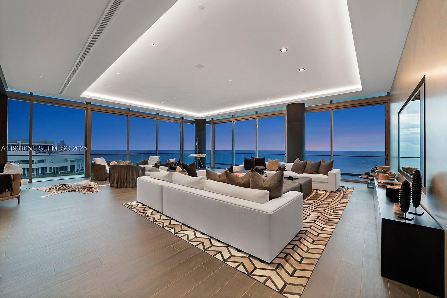 This is a luxurious living room featuring floor-to-ceiling windows with ocean views. The room is furnished with a large white sectional sofa, decorative pillows, and a geometric patterned rug. A modern entertainment center and stylish decor enhance the sophisticated ambiance of the space.