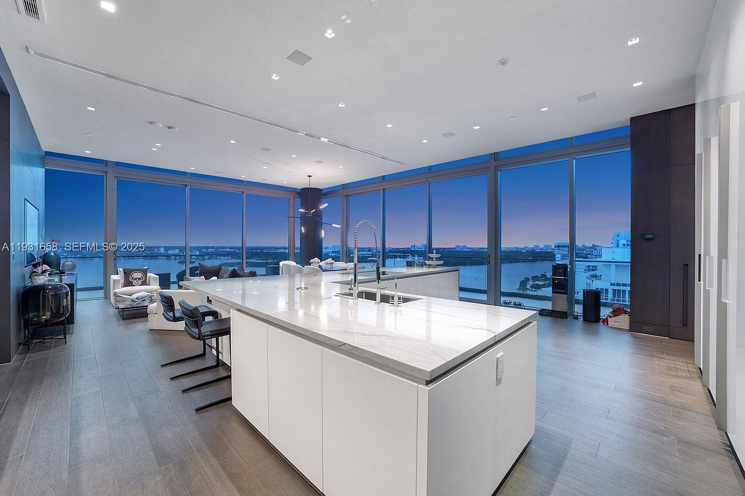This is a modern, high-end kitchen featuring a large island with a white marble countertop and white cabinetry. The kitchen boasts floor-to-ceiling windows offering a panoramic view of the city and waterfront. The open-concept design flows into a living area, creating a spacious and luxurious atmosphere.
