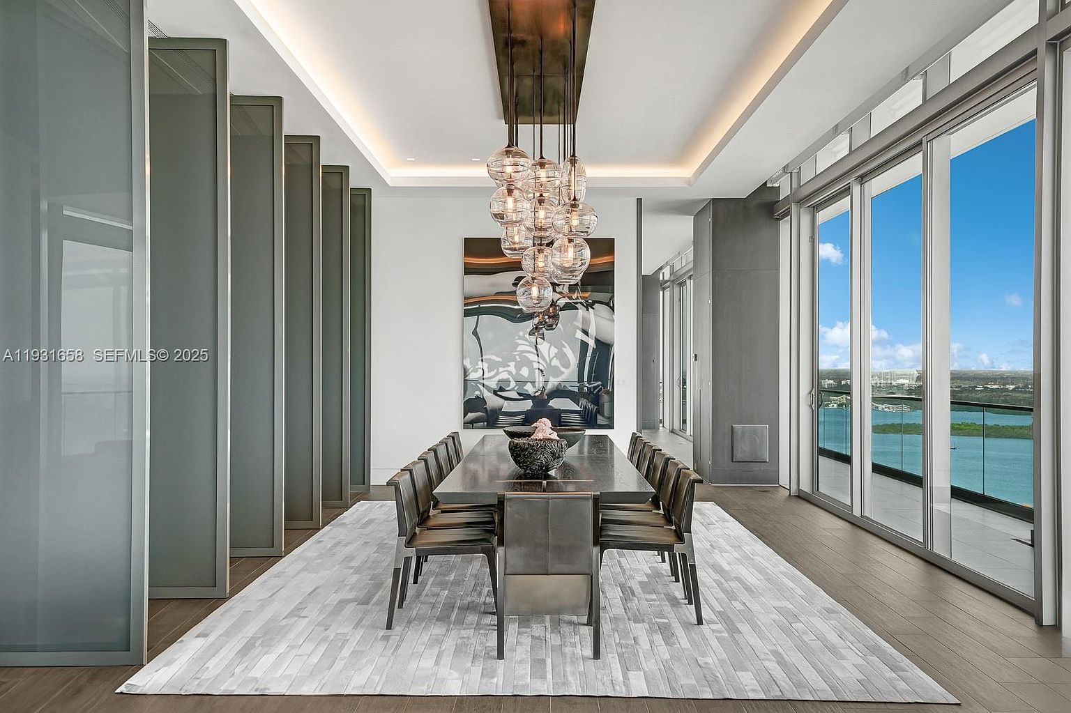 This is a modern dining room featuring a long, dark wood table with seating for ten, illuminated by a unique cluster of glass pendant lights. A large abstract painting hangs on the wall, and floor-to-ceiling windows offer a stunning view of the water. The room is decorated in neutral tones with a light-colored rug, creating a sophisticated and elegant atmosphere.