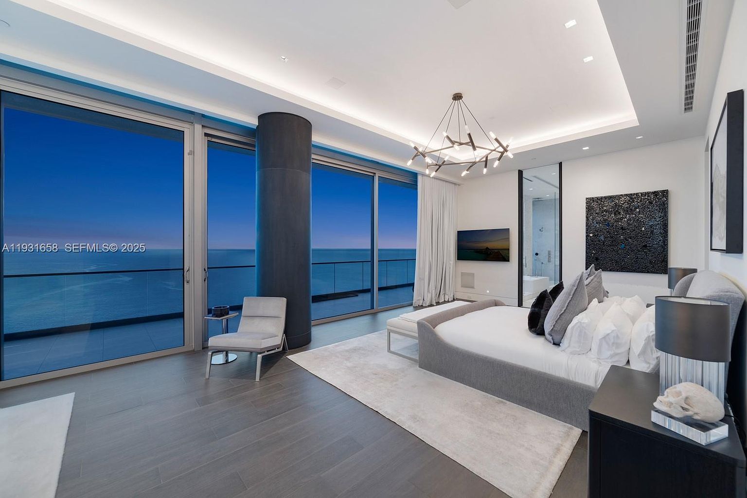 This is a luxurious primary bedroom featuring floor-to-ceiling windows with ocean views, a modern chandelier, and a unique bed design. The room is decorated in a neutral color palette with gray wood floors, creating a serene and sophisticated atmosphere. A modern art piece hangs above the bed, adding a touch of elegance.
