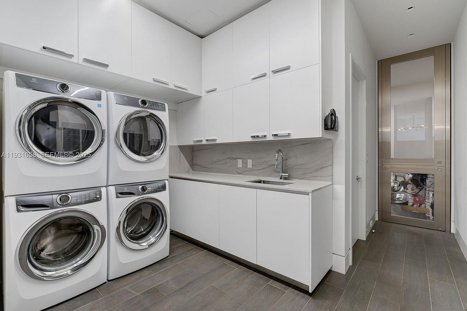 This is a well-organized laundry room featuring stacked washer and dryer units, white cabinetry with sleek hardware, and a countertop with a sink. The room has a modern aesthetic with gray wood-look flooring and a marble backsplash. A doorway leads to another room, possibly a pantry or storage area.