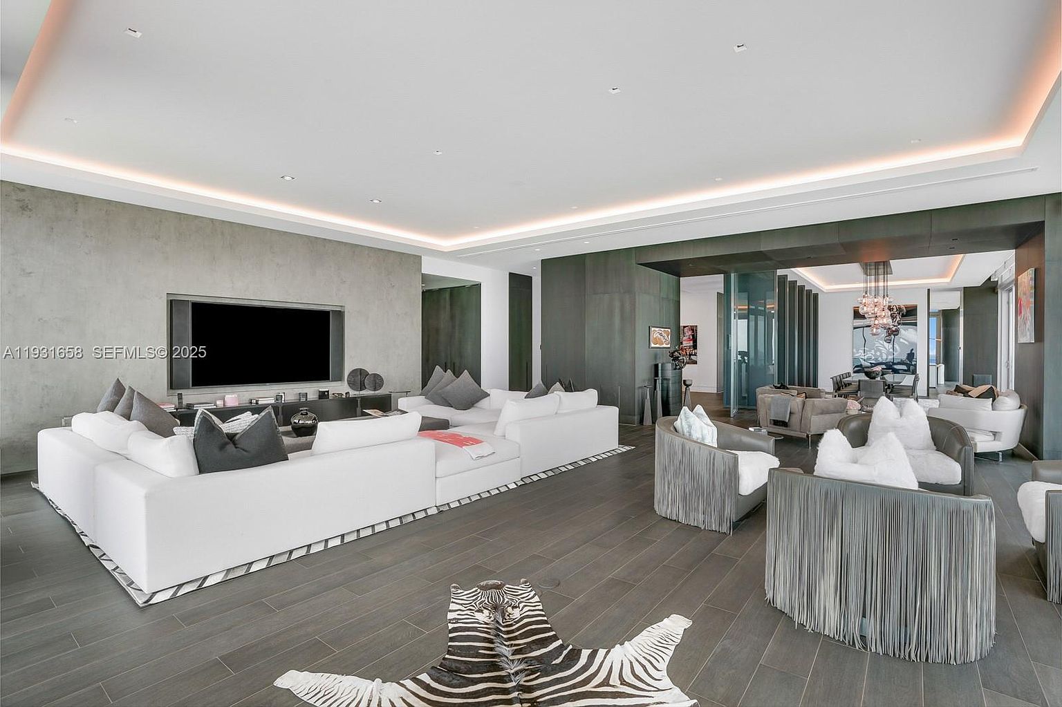 This is a spacious and modern living room featuring a large white sectional sofa, a zebra-print rug, and stylish armchairs with fringe detailing. A large flat-screen TV is mounted on a textured wall, and the room opens into a dining area with a chandelier. The overall impression is luxurious and contemporary.