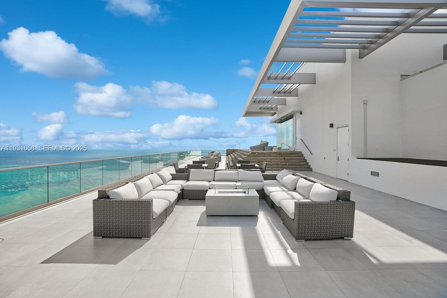 This image showcases a luxurious outdoor patio or balcony area with stunning ocean views. A large, modular seating arrangement with white cushions surrounds a modern fire pit, creating an inviting space for relaxation and entertainment. The sleek, contemporary design is complemented by the clean lines of the building's architecture and the expansive blue sky.