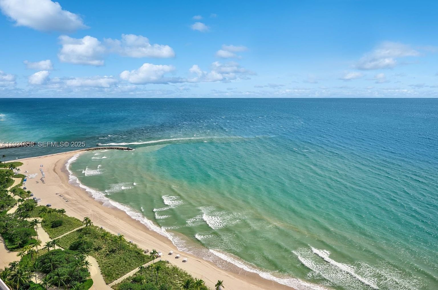 This aerial view showcases a stunning coastline with turquoise waters meeting a sandy beach. Lush greenery lines the shore, providing a vibrant contrast to the ocean. A pier extends into the water, adding a focal point to the expansive seascape under a partly cloudy sky.