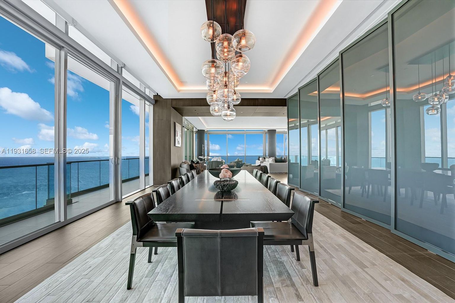 This is a luxurious dining room featuring a long, dark wood table with numerous chairs, illuminated by a modern chandelier with multiple glass orbs. Large windows offer stunning ocean views, enhancing the room's appeal. The space is designed with a contemporary aesthetic, emphasizing elegance and sophistication, making it ideal for upscale dining and entertaining.