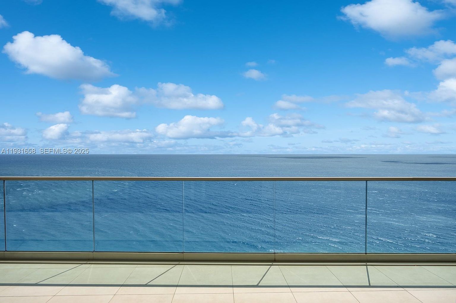 This image showcases a stunning ocean view from a balcony. The balcony features a glass railing that provides an unobstructed view of the water and sky. The tile flooring is neutral and clean, and the overall impression is one of luxury and tranquility.