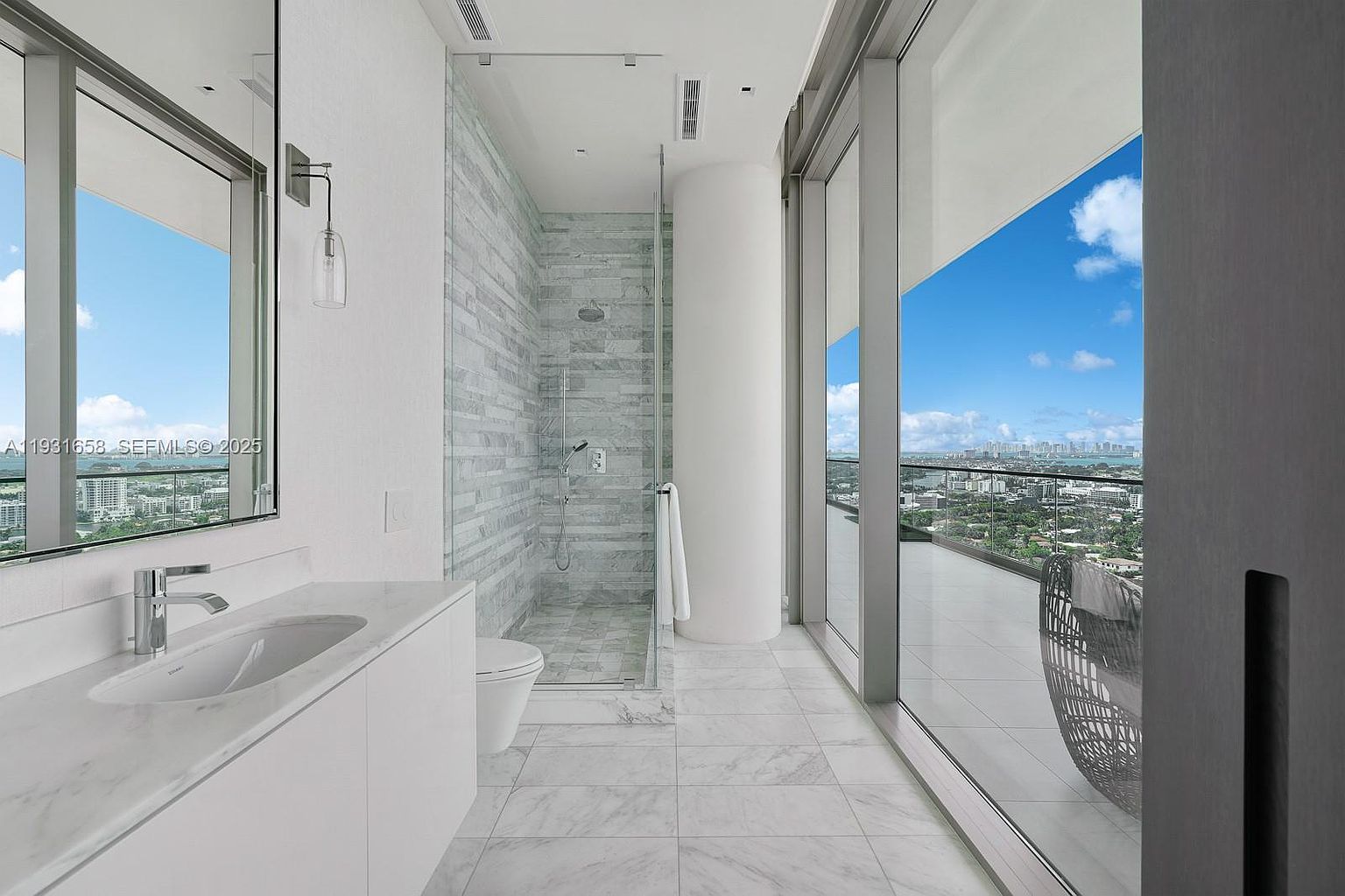 This is a modern primary bathroom featuring a sleek vanity with a white countertop and integrated sink, a glass-enclosed shower with marble tile, and a toilet. Large windows offer expansive views of the city skyline and a balcony, creating a bright and airy atmosphere. The overall design is minimalist and luxurious.