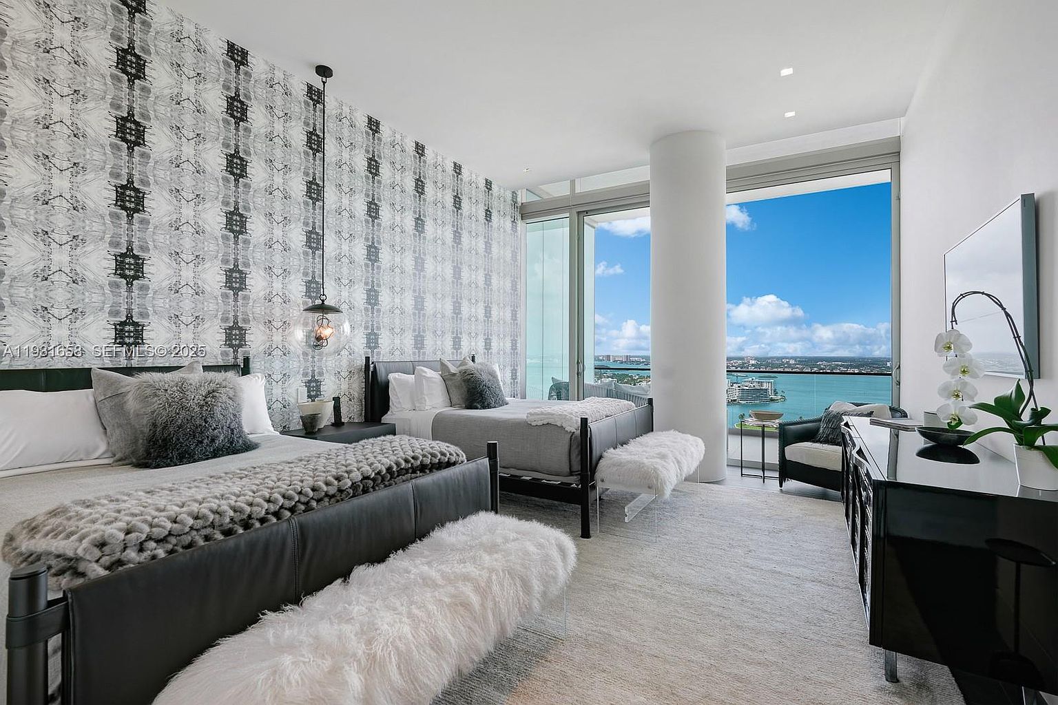 This is a luxurious primary bedroom featuring two beds with dark frames and plush bedding. The room has a modern design with patterned wallpaper, a statement pendant light, and floor-to-ceiling windows offering a stunning water view. A sleek black dresser and a comfortable armchair add to the room's elegance.