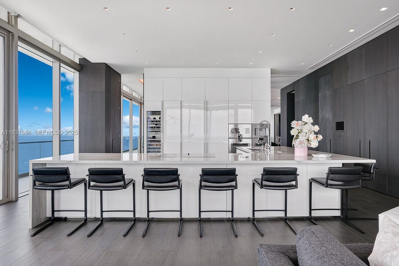 This is a modern, high-end kitchen featuring a large island with seating, sleek white cabinetry, and dark wood accents. The kitchen is well-lit with natural light from a large window offering a view of the ocean. The overall impression is one of luxury and sophistication.