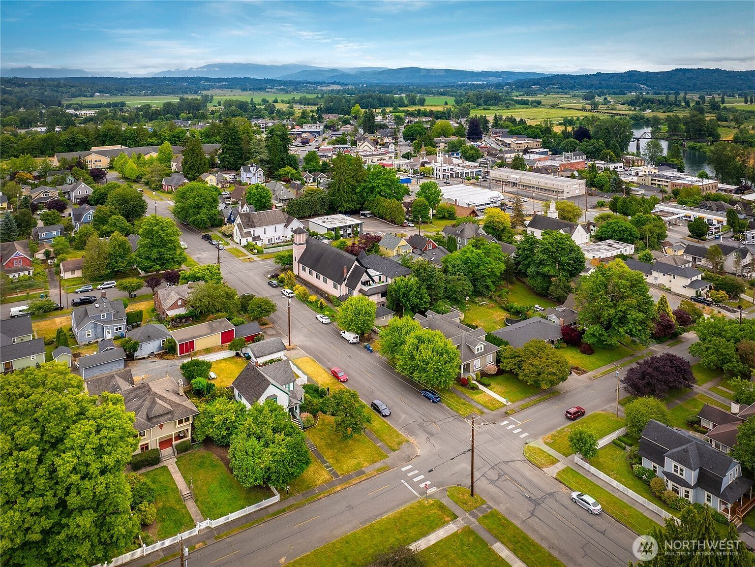This aerial view showcases a charming residential neighborhood with a mix of architectural styles, lush green trees, and well-maintained lawns. The town center is visible in the distance, featuring a mix of commercial and civic buildings. The overall impression is one of a peaceful and well-established community.