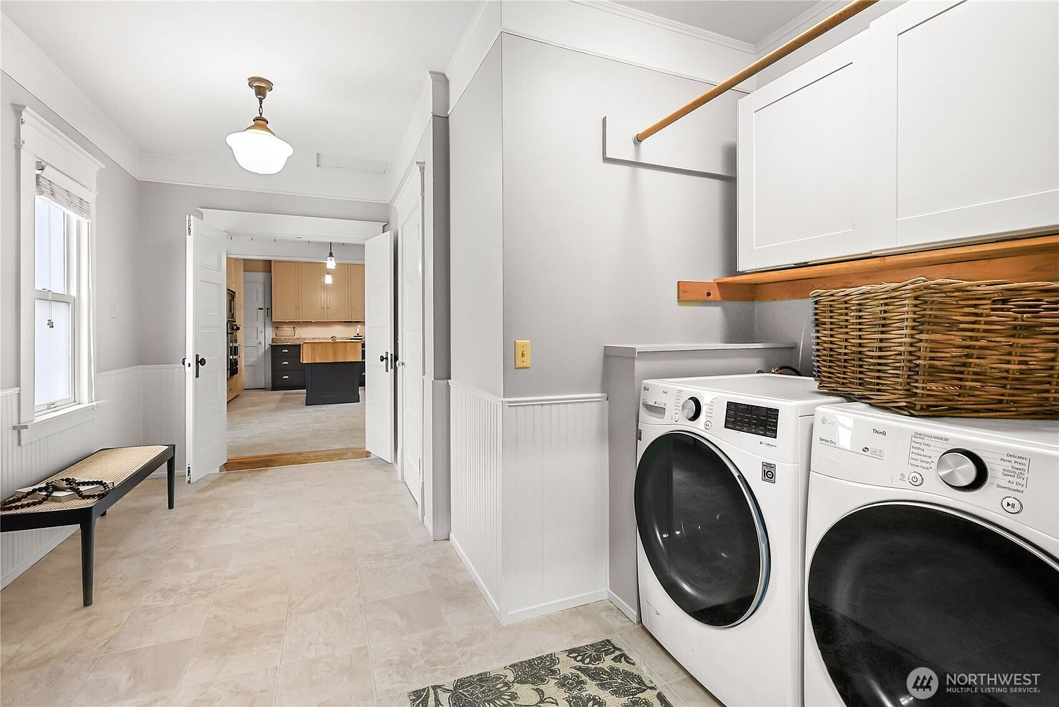 This is an interior shot of a laundry room featuring a washer and dryer set, white cabinetry, and a wooden hanging rod. The room has a neutral color palette with light gray walls and beige tile flooring. A bench is visible in the adjacent hallway, leading to a kitchen area, suggesting a functional and well-connected layout.