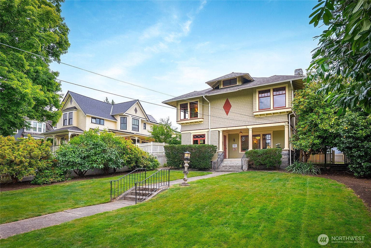 This is a front exterior view of a well-maintained two-story house with a green lawn. The house features a covered porch with columns, a diamond-shaped decorative element, and a chimney. A walkway leads to the entrance, and there's a second house visible in the background, suggesting a residential neighborhood.