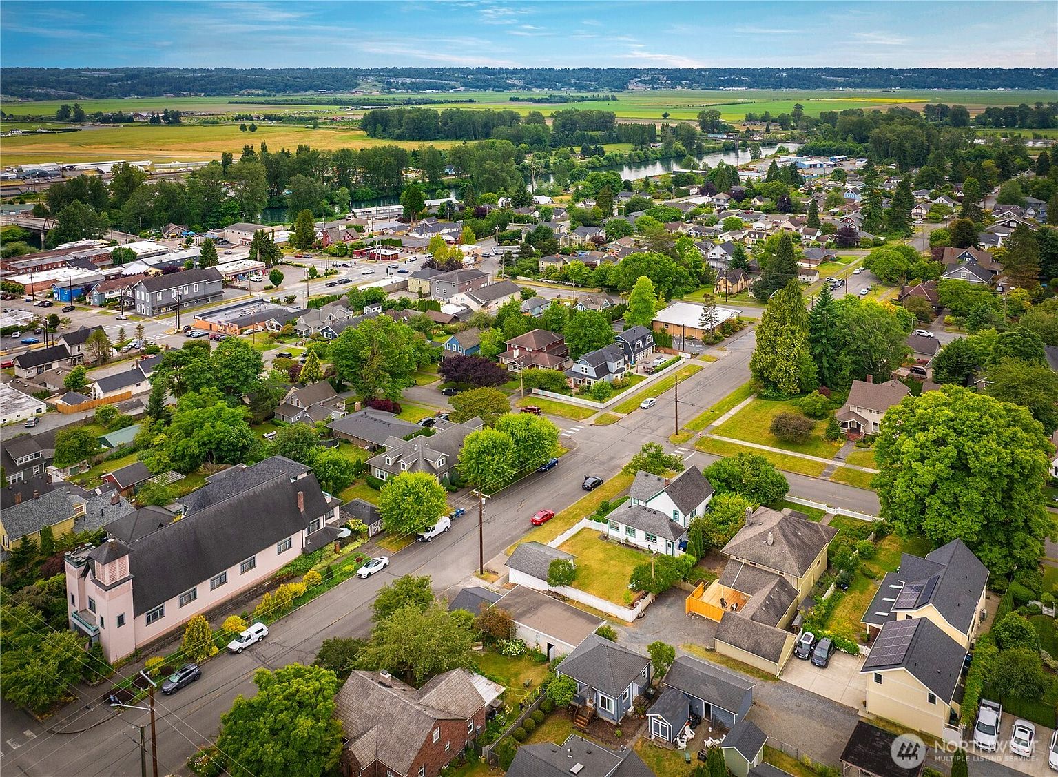 This aerial view showcases a charming residential neighborhood with a mix of single-family homes, mature trees, and well-maintained streets. The houses feature various architectural styles, and the landscape includes green lawns and gardens. In the background, there are open fields and a river, adding to the scenic appeal of the area.