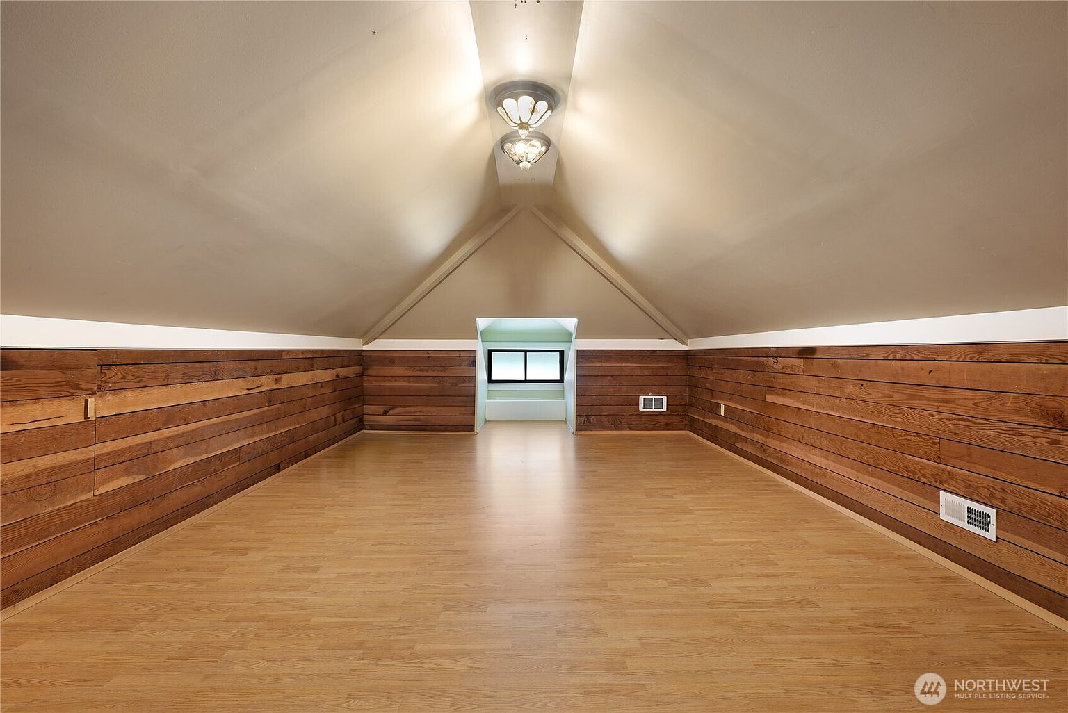 This is an interior shot of an attic space, featuring wood-paneled walls and light-colored hardwood flooring. The room has a distinctive A-frame ceiling with two light fixtures. A small window area is visible at the far end of the room, providing natural light and a view to the outside.