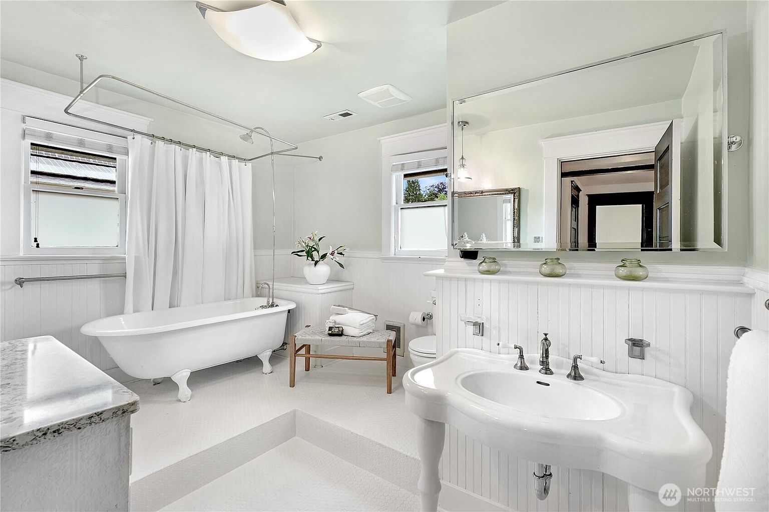 This is a bright and airy primary bathroom featuring a clawfoot tub with a shower curtain, a pedestal sink, and white wainscoting. A large mirror reflects the room, enhancing the sense of space. The overall style is classic and elegant, creating a relaxing atmosphere.
