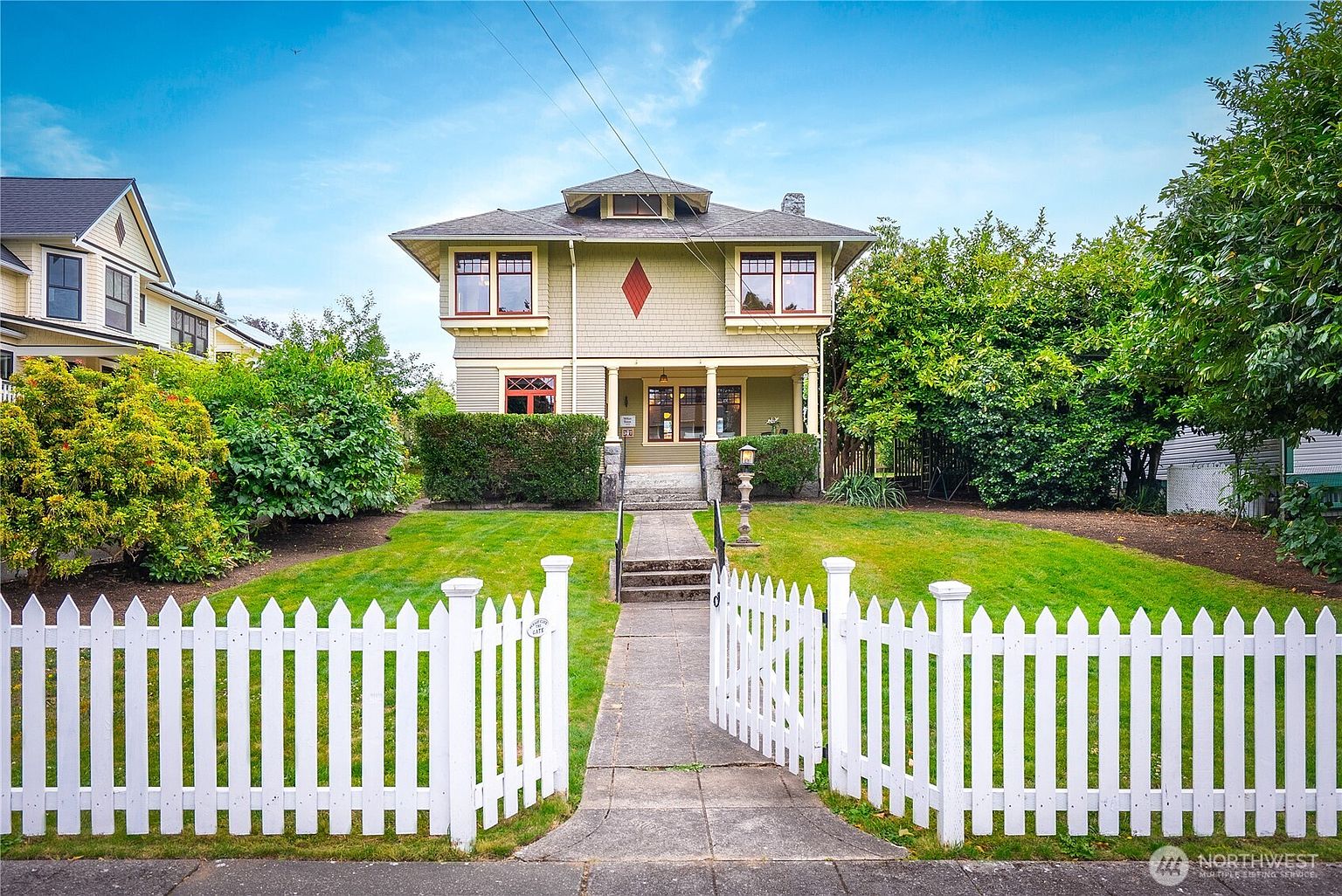 This is a charming front view of a two-story house with a well-manicured lawn and a white picket fence. The house features a symmetrical design with a diamond-shaped detail on the second floor, complemented by red-framed windows and a covered porch. The overall impression is one of classic American suburban charm and curb appeal.