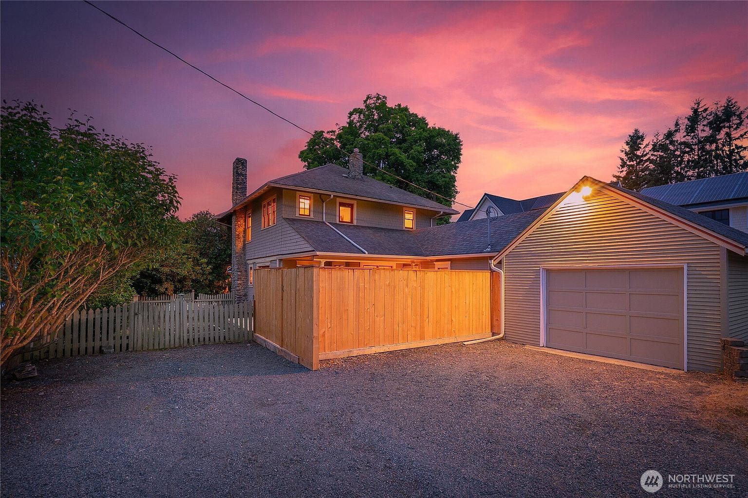 The image showcases the rear exterior of a charming two-story house with a detached garage. A wooden fence encloses part of the backyard, providing privacy. The gravel driveway leads to the garage, and the sky is painted with vibrant hues of pink and purple during sunset, creating a warm and inviting atmosphere.