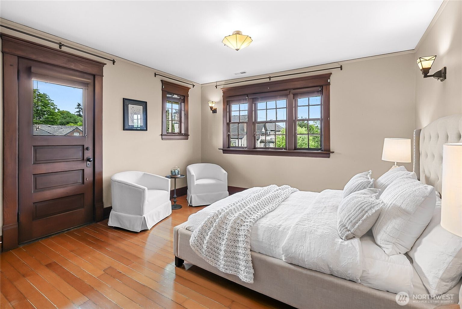 This is a primary bedroom featuring hardwood floors, a neutral color palette, and dark wood trim around the windows and door. The room is furnished with a large bed with a tufted headboard, white bedding, and two white armchairs. Natural light streams in through the windows, creating a bright and inviting space.
