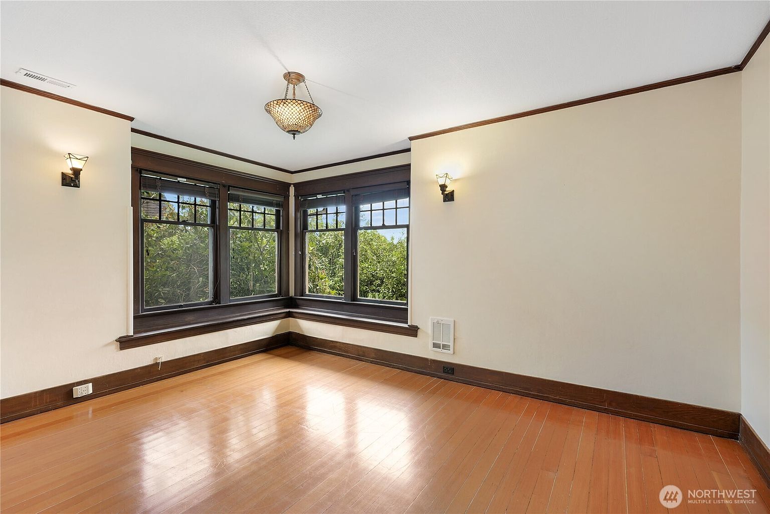 This is an interior shot of a living room featuring hardwood floors and dark wood trim around the windows and ceiling. The room has a vintage charm with a decorative light fixture and wall sconces. Large windows offer a view of lush greenery outside, creating a bright and inviting space.