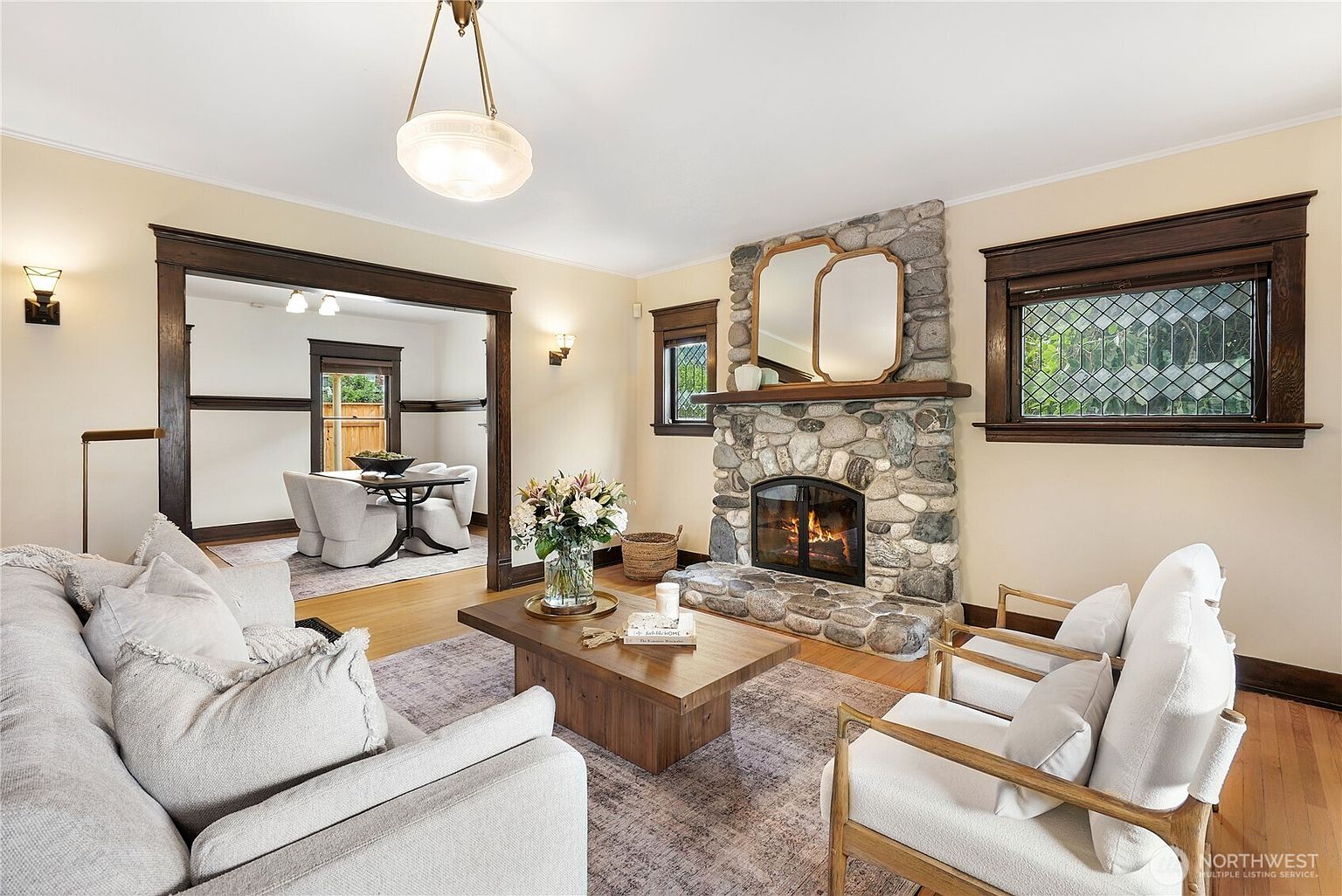 This is an inviting living room featuring a stone fireplace with a wooden mantel and a decorative mirror. The room is furnished with a comfortable light-colored sofa, two armchairs, and a wooden coffee table adorned with flowers and books. Natural light streams in through the windows, highlighting the hardwood floors and creating a warm, cozy atmosphere.