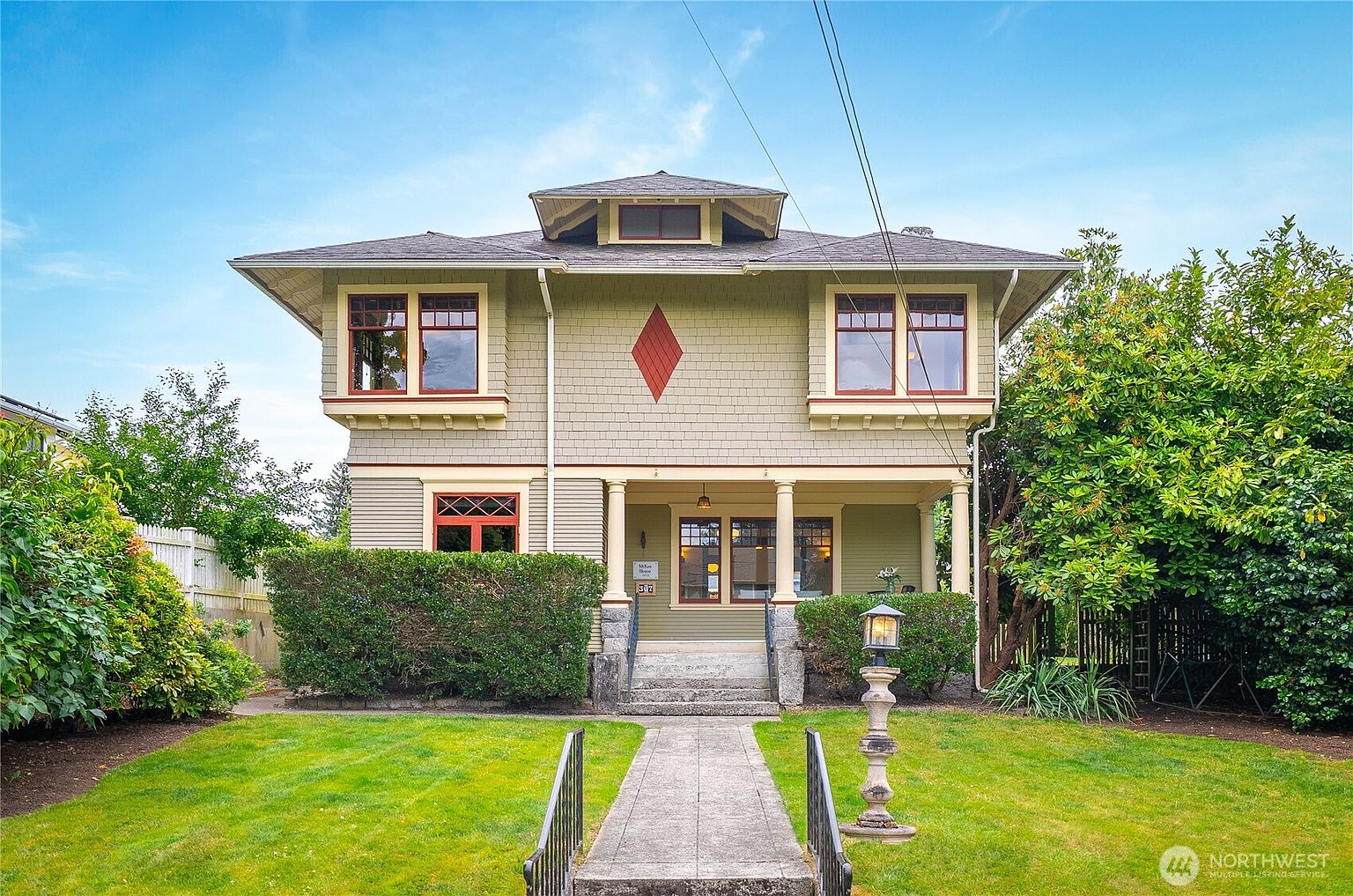 The image showcases the front exterior of a two-story craftsman-style home with a well-manicured lawn and mature landscaping. The house features a covered porch supported by columns, multiple windows with red trim, and a distinctive diamond-shaped detail on the facade. A concrete walkway leads to the entrance, flanked by a low hedge and a vintage-style lamppost, creating a welcoming and charming curb appeal.