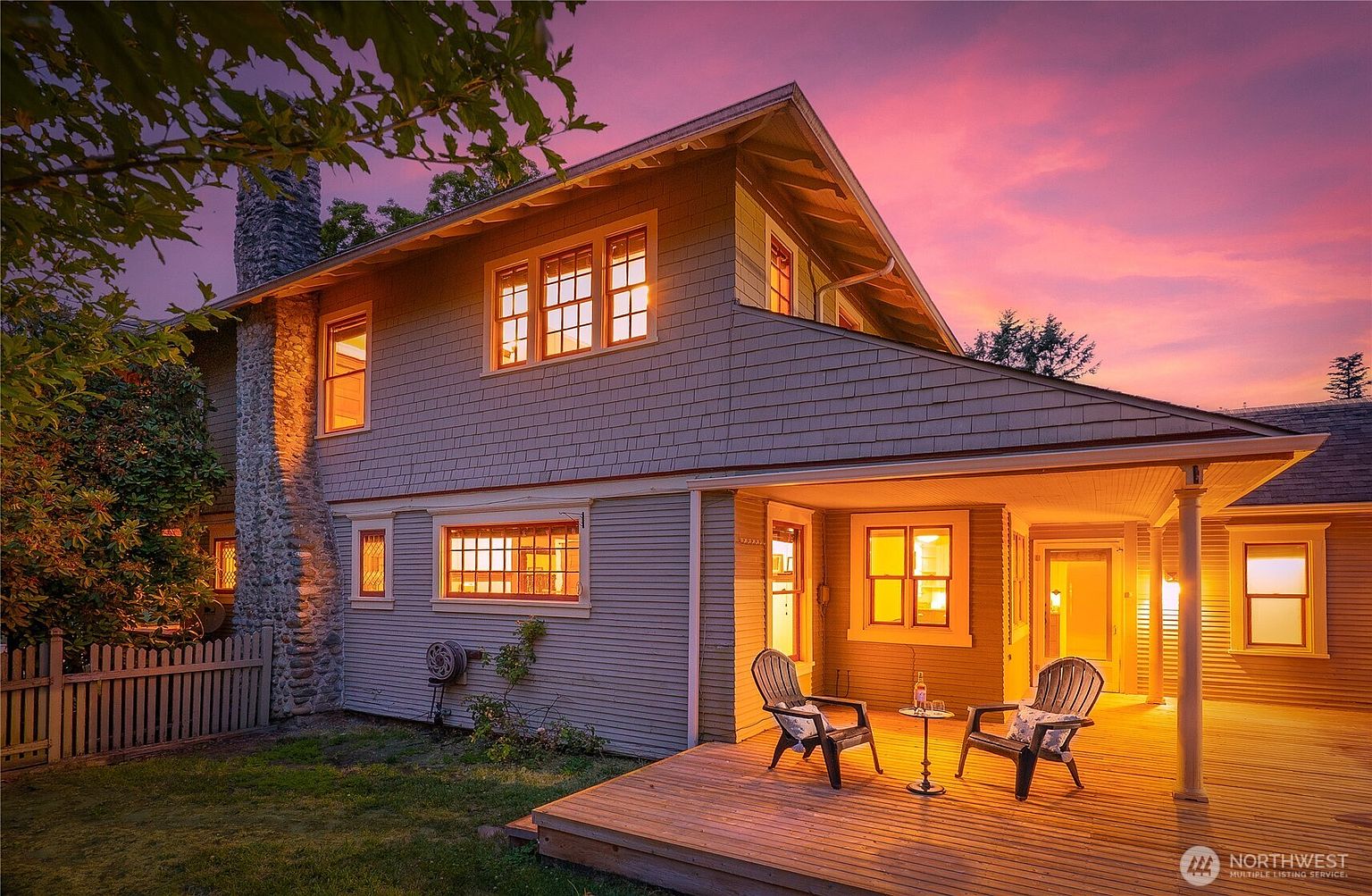 This image showcases a charming home exterior at dusk, featuring a cozy patio with two chairs and a small table set for relaxation. The warm glow from the house's windows contrasts beautifully with the vibrant pink and purple sky, creating an inviting and serene atmosphere. The well-maintained wooden deck and the surrounding lawn add to the property's appeal.