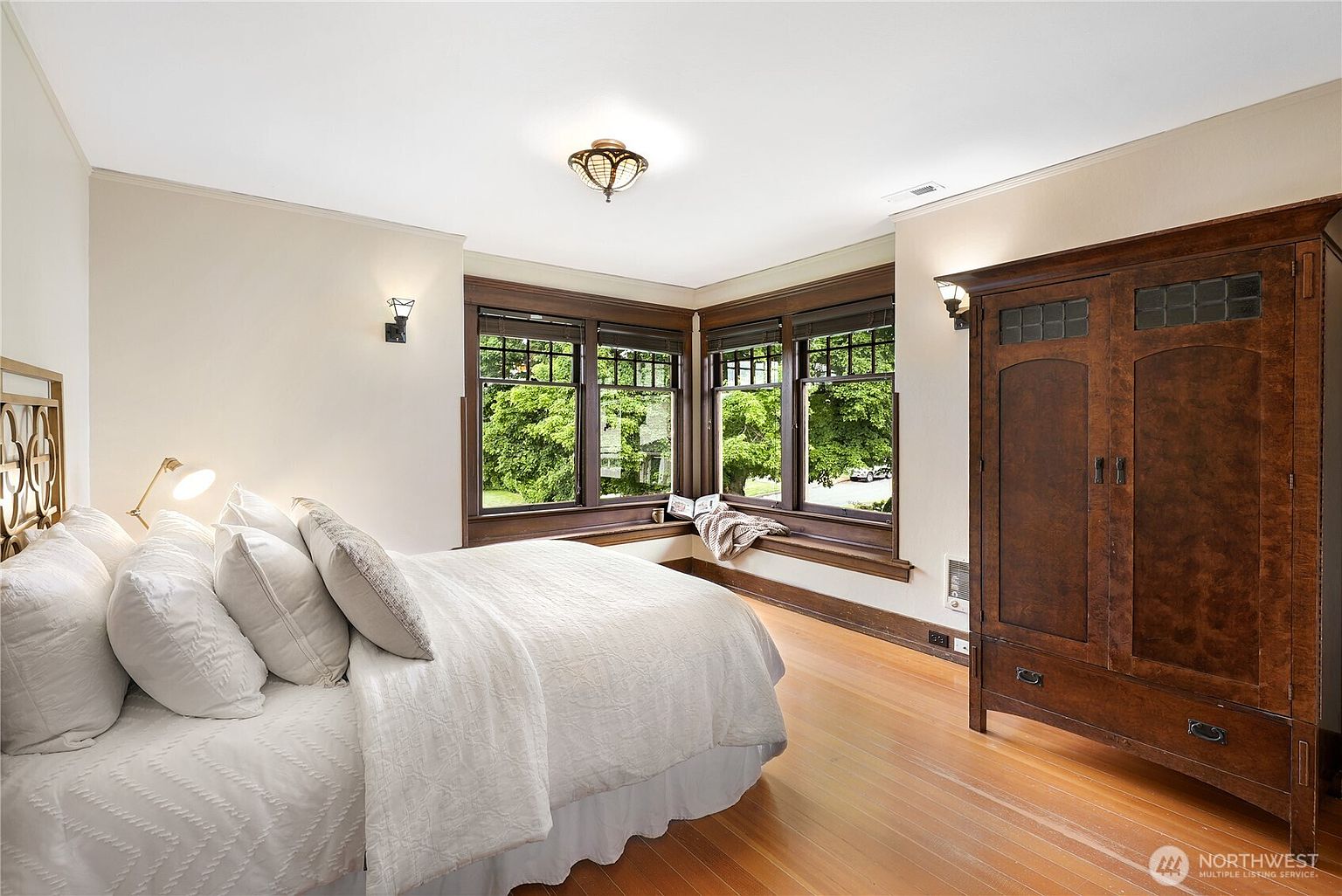 This is a cozy primary bedroom featuring a large bed with white linens and pillows, a bay window with dark wood trim, and a matching dark wood wardrobe. The room has hardwood floors and neutral-colored walls, creating a warm and inviting atmosphere. The lighting fixtures add a touch of vintage charm.