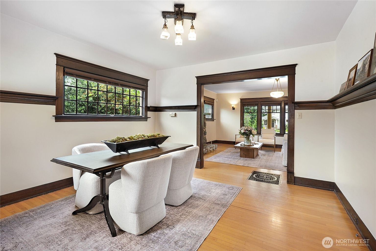 This is an interior shot of a dining room featuring a rectangular table with a dark wood frame and a light-colored top, surrounded by four upholstered chairs. The room has dark wood trim around the window and along the walls, contrasting with the light-colored walls. A doorway leads into another room, suggesting an open floor plan, and a patterned rug lies beneath the dining set, adding texture to the hardwood floor.
