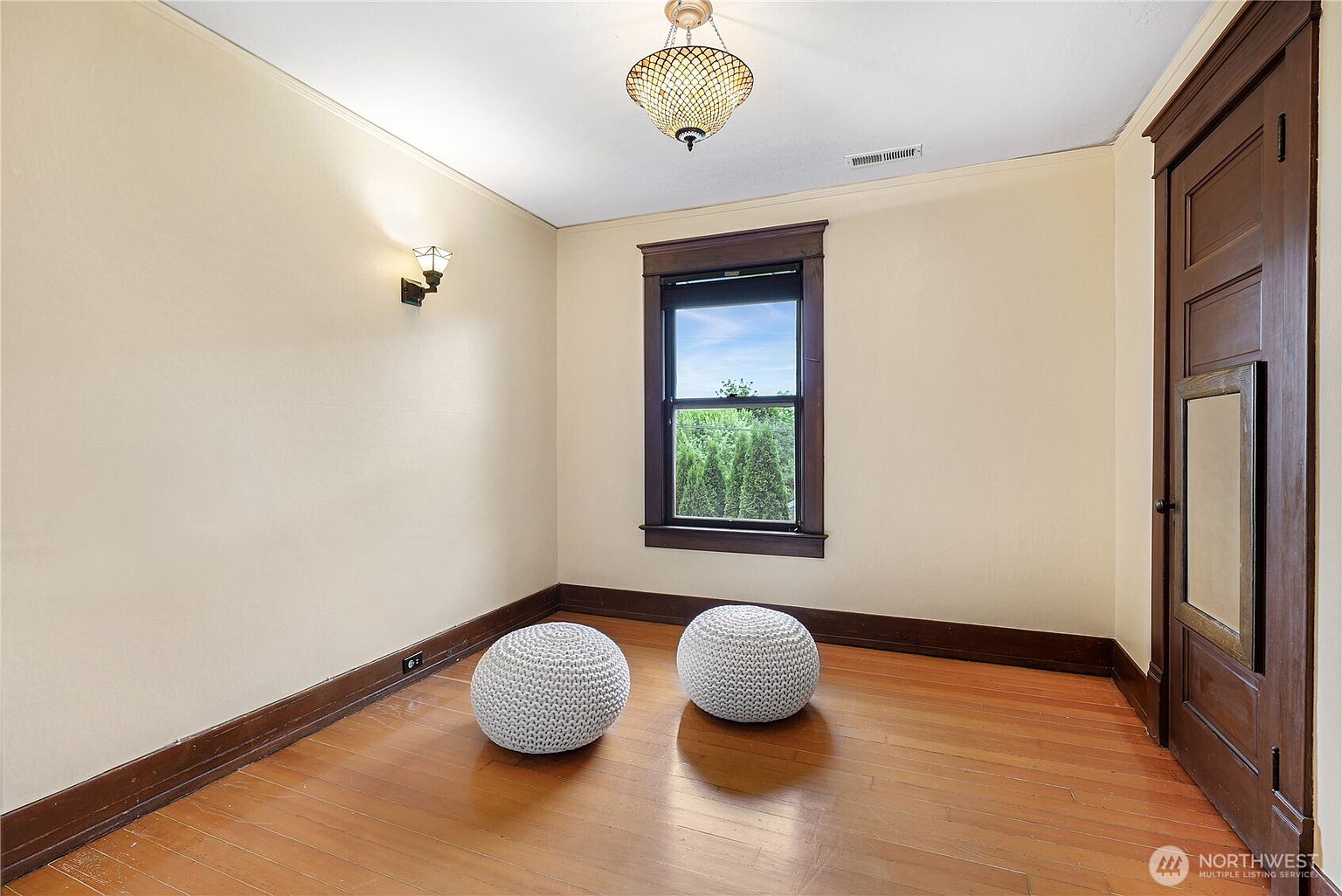 The image showcases a well-lit bedroom with hardwood floors and neutral-toned walls. Two round, knitted poufs sit on the floor, adding a touch of casual comfort. A window with dark trim provides natural light, and a vintage-style chandelier hangs from the ceiling, complementing the dark wood trim around the room and door.