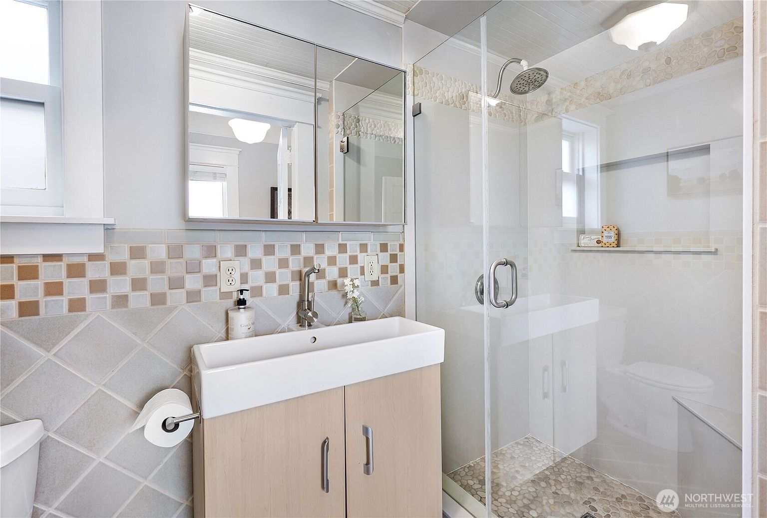 This is a well-lit bathroom featuring a modern vanity with a rectangular sink and light wood cabinetry. A mirrored medicine cabinet hangs above the sink, and the walls are adorned with a combination of square and diamond-patterned tiles. A glass-enclosed shower with pebble flooring adds a touch of luxury.