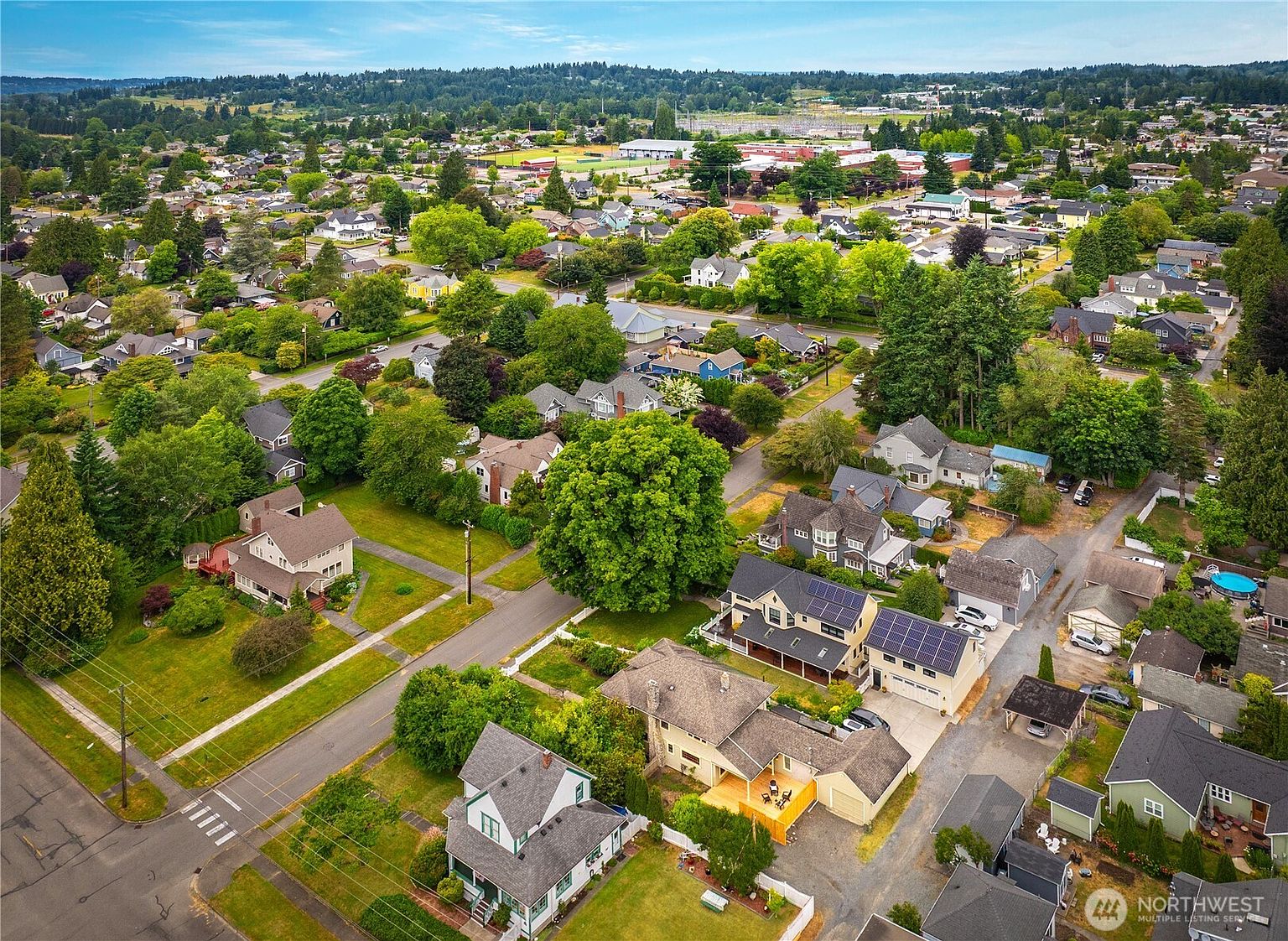 This aerial view showcases a well-maintained residential neighborhood with lush green trees and manicured lawns. Several houses feature solar panels, and the streets are clean and orderly. In the distance, a school or community center is visible, adding to the appeal of the location.