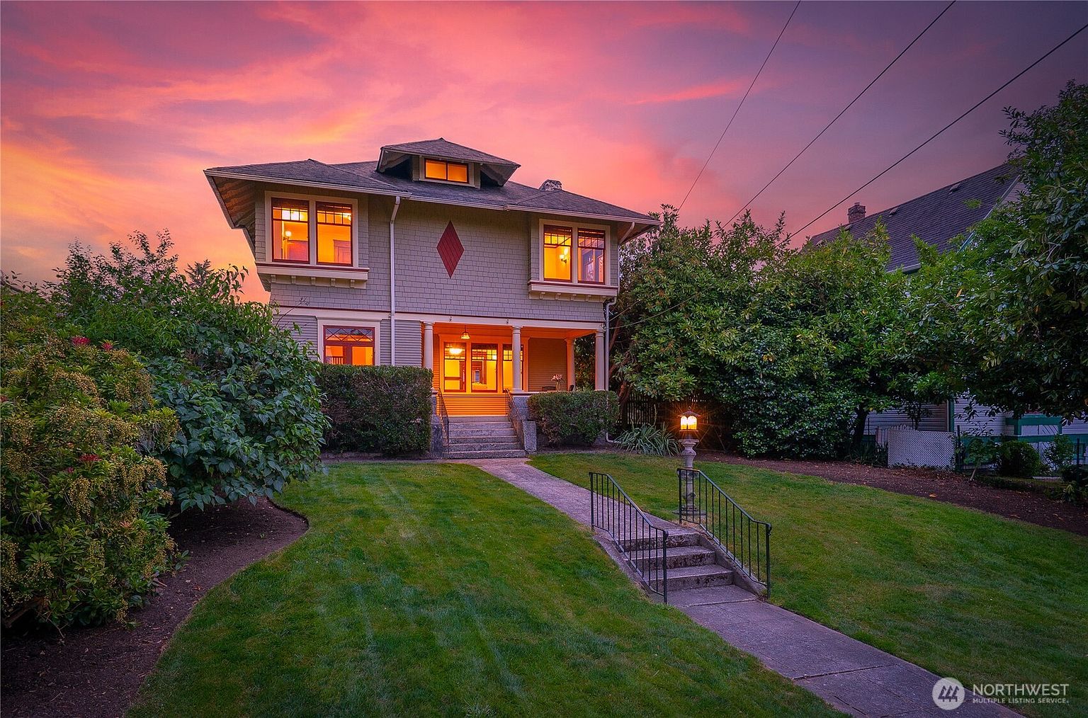 This is a captivating front view of a two-story house at dusk, showcasing its charming curb appeal. The well-manicured lawn leads to a welcoming entryway with steps and a railing, while the house itself features a symmetrical design with illuminated windows and a distinctive diamond-shaped detail. The warm glow emanating from the windows contrasts beautifully with the vibrant colors of the twilight sky, creating an inviting and picturesque scene.