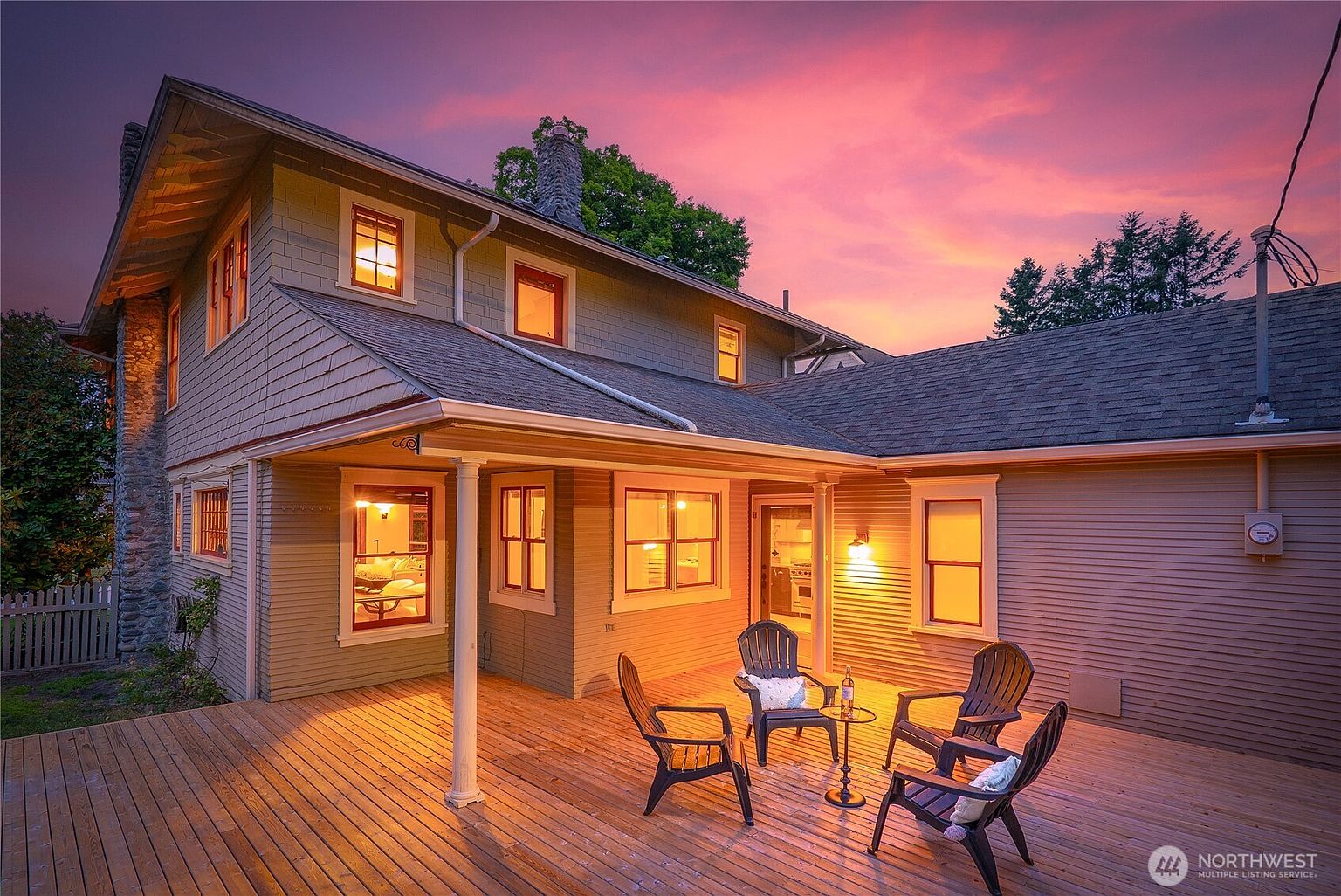 This image showcases a charming outdoor deck area at dusk, featuring wooden flooring and comfortable seating arrangements. The warm glow from the house's windows creates an inviting atmosphere, complemented by the colorful sunset sky. The scene suggests a perfect spot for relaxation and outdoor entertainment.