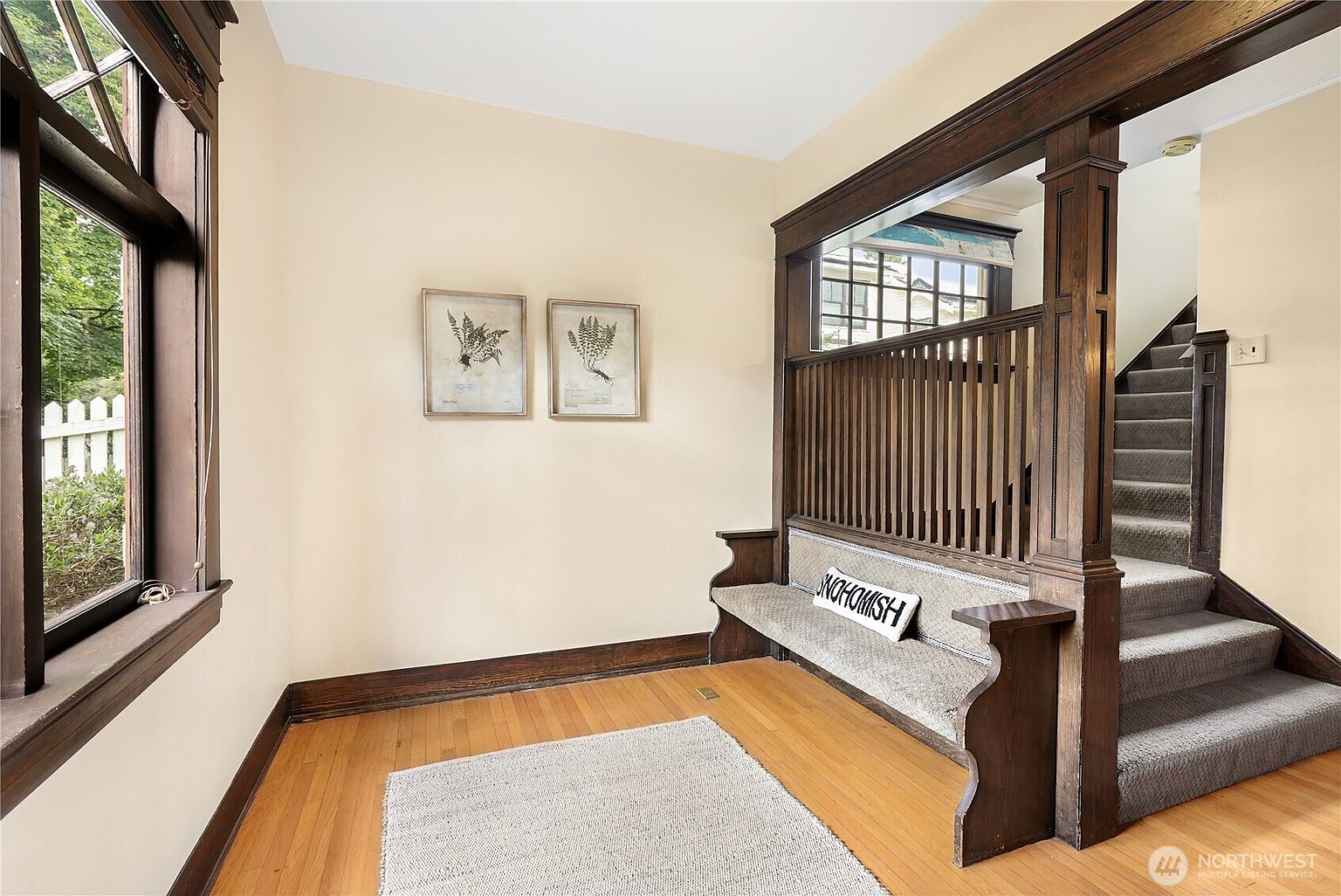 This interior shot showcases a charming hallway with a built-in bench and staircase. The dark wood trim contrasts beautifully with the light walls and hardwood floors, creating a warm and inviting atmosphere. Two framed botanical prints add a touch of elegance to the space.