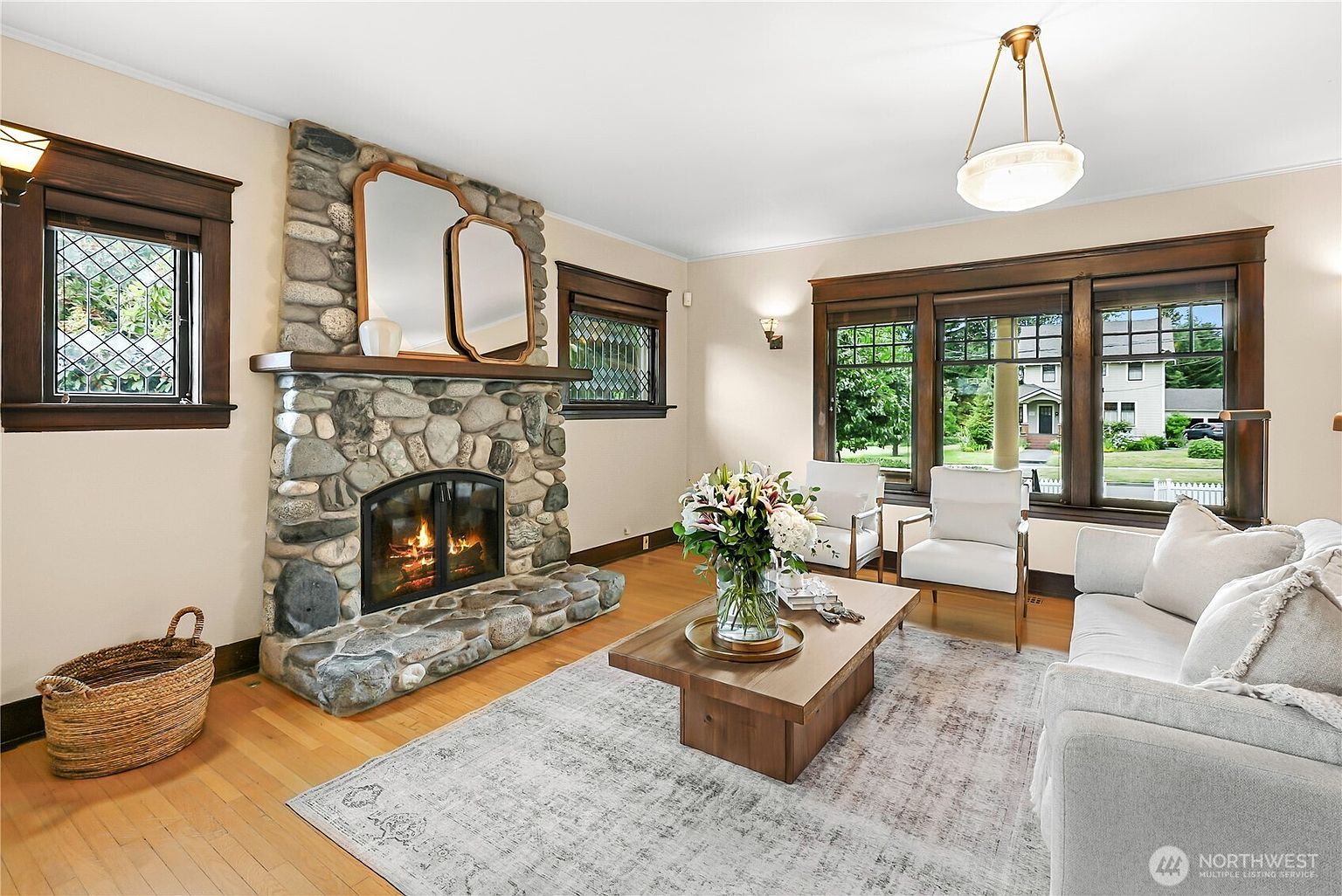This is a cozy living room featuring a stone fireplace with a wooden mantel and two decorative mirrors. The room is furnished with a light-colored sofa, two white armchairs, and a wooden coffee table adorned with a floral arrangement. Natural light streams in through the windows, highlighting the hardwood floors and the neutral color palette of the walls and area rug.
