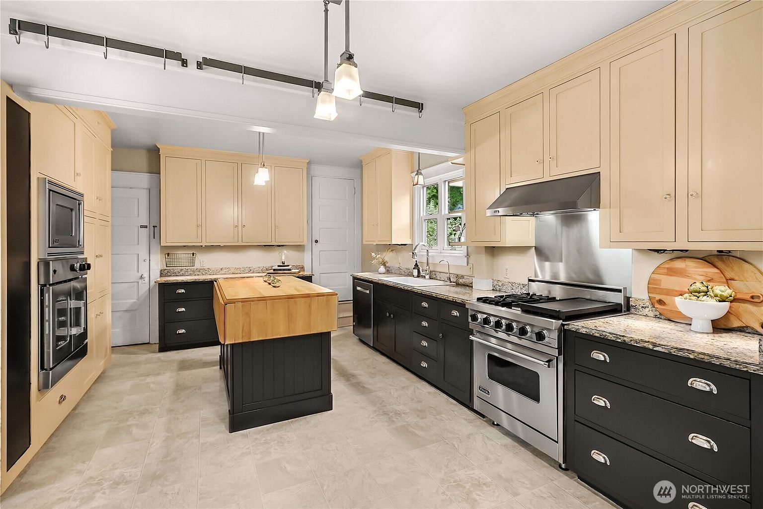 This is a well-lit kitchen featuring a mix of light and dark cabinetry, granite countertops, and stainless steel appliances. A central island with a butcher block top provides additional workspace. The kitchen has a traditional style with modern touches, creating a warm and inviting atmosphere.