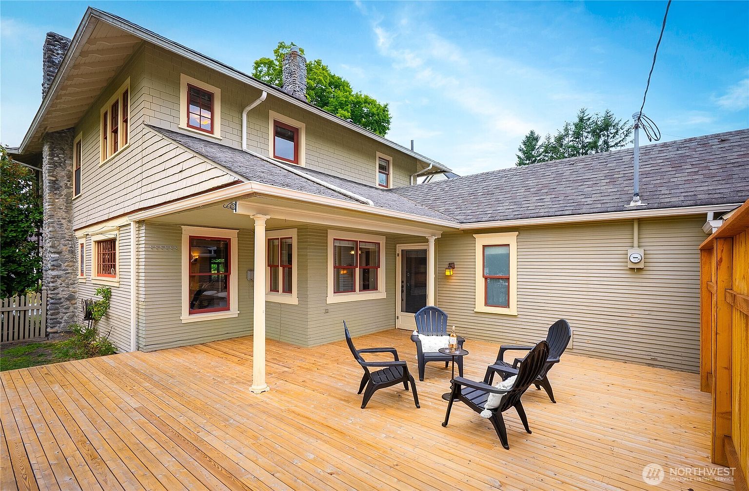 This image showcases a spacious wooden deck area attached to a charming two-story house. The deck is furnished with several black chairs arranged around a small table, creating an inviting outdoor living space. The house features a mix of siding and stone elements, with red-framed windows adding a pop of color against the muted green exterior.