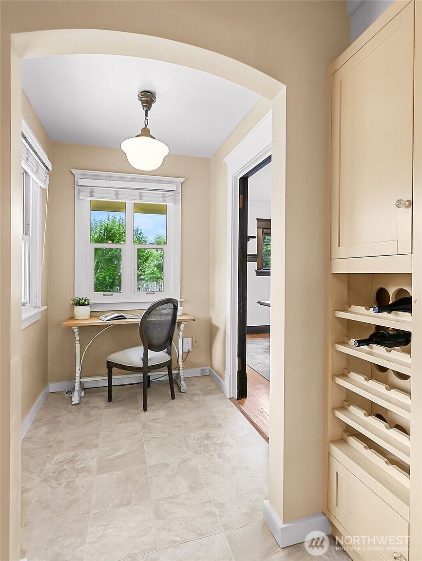 This is an interior shot of a small office or study area. The room features a desk with a chair, a window providing natural light, and built-in wine storage. The color palette is neutral, and the overall impression is cozy and functional.