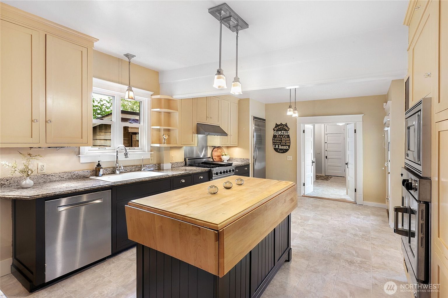 This is a well-lit kitchen featuring a mix of light and dark cabinetry, granite countertops, and stainless steel appliances. A central island with a butcher block top provides ample workspace, while pendant lighting adds a touch of elegance. The kitchen opens to a hallway, creating a sense of flow and connectivity within the home.