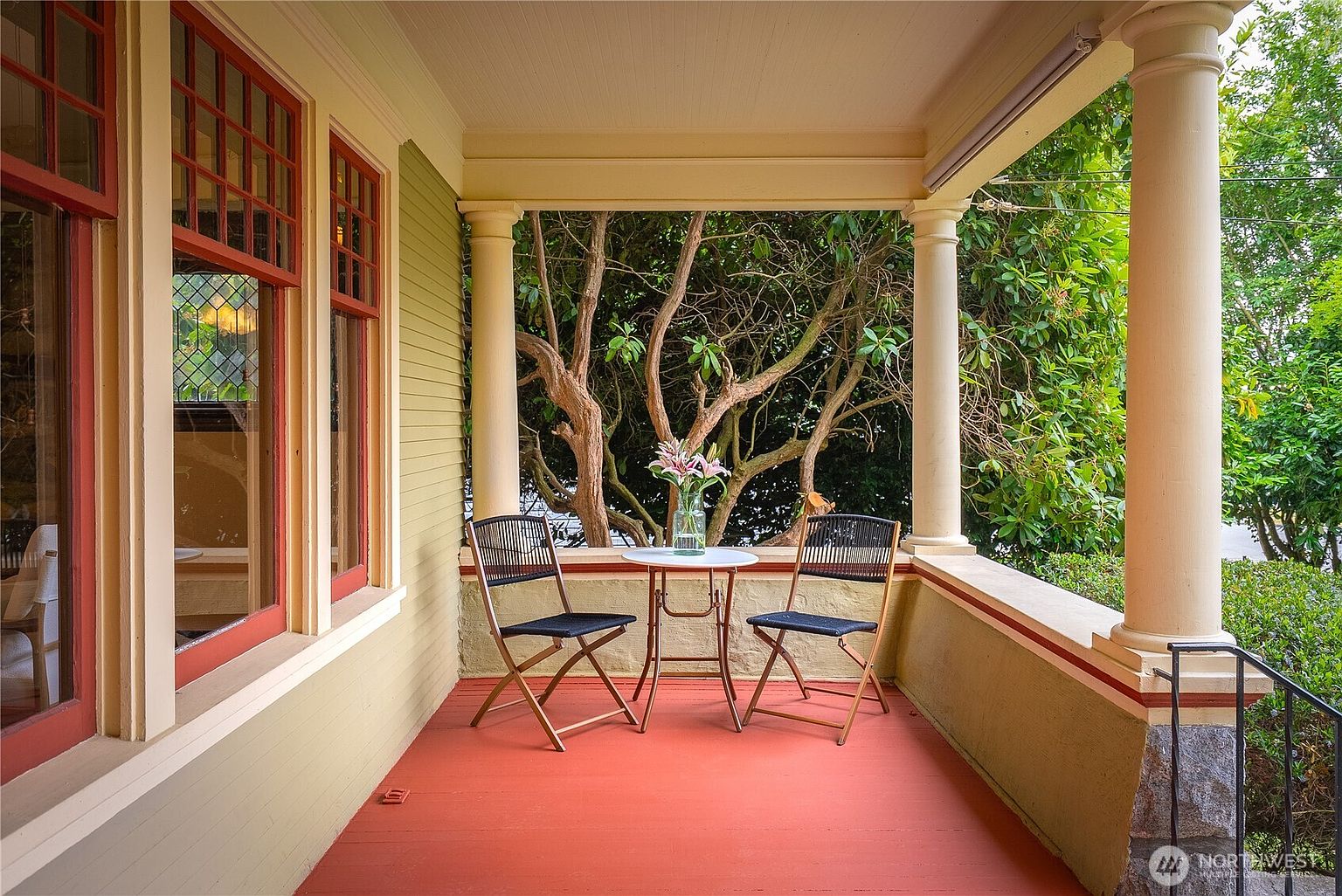 This image showcases a charming outdoor patio or balcony area. Two chairs and a small table with a vase of flowers create an inviting seating arrangement. The space is framed by architectural columns and offers a view of lush greenery, suggesting a peaceful and private setting.