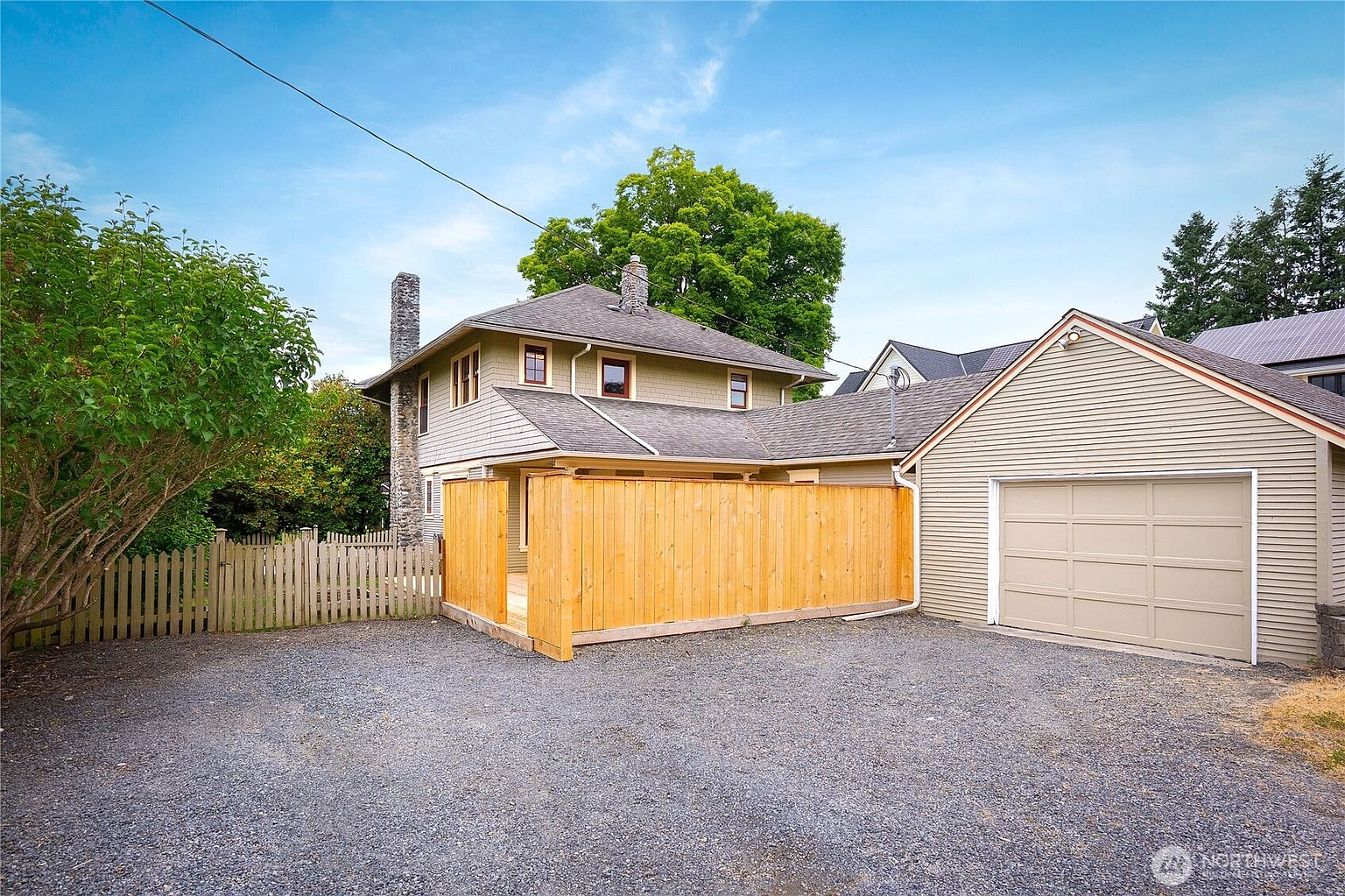 This image showcases the rear exterior of a charming home, featuring a combination of shingle and siding. A wooden fence encloses a portion of the backyard, providing privacy. A detached garage is visible on the right, and the gravel driveway offers ample parking space.