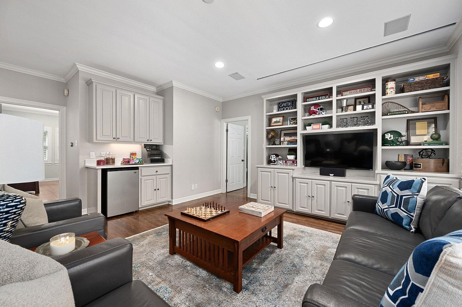 This living room features a built-in shelving unit with sports memorabilia and a television. A dark leather sofa and armchairs are arranged around a wooden coffee table with a chess set. The room has a cozy and inviting atmosphere, perfect for relaxation and entertainment.