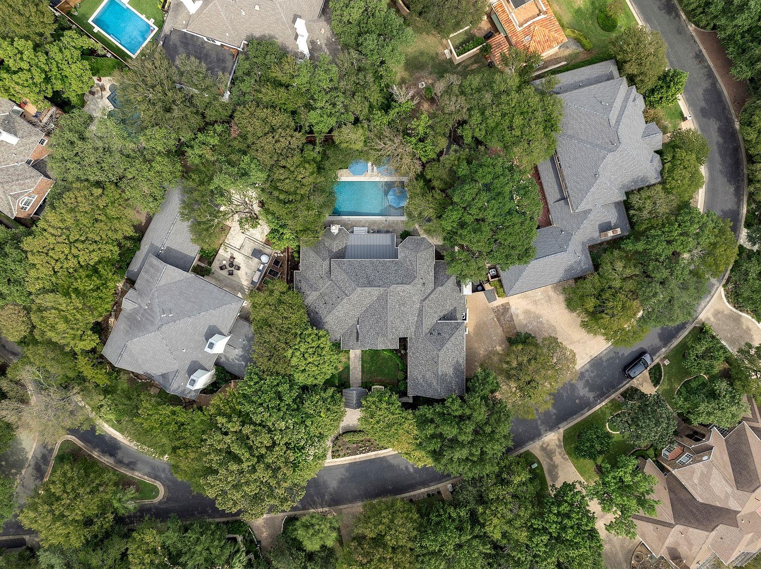 This aerial view showcases a luxurious property nestled among mature trees, highlighting its privacy and serene setting. The house features a complex roofline, a swimming pool, and a circular driveway, suggesting a high-end, secluded estate. The surrounding landscaping and neighborhood layout emphasize exclusivity and spaciousness.