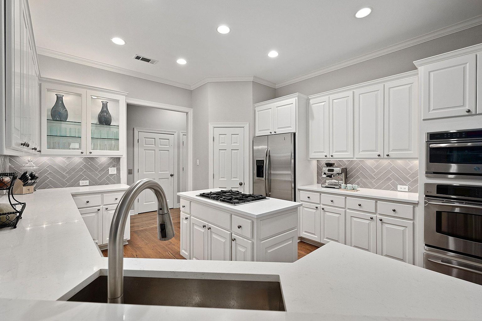 This is a bright and modern kitchen featuring white cabinetry, stainless steel appliances, and hardwood floors. A central island with a gas cooktop provides ample workspace, while the herringbone backsplash adds a touch of elegance. The kitchen is well-lit with recessed lighting, creating a clean and inviting atmosphere.