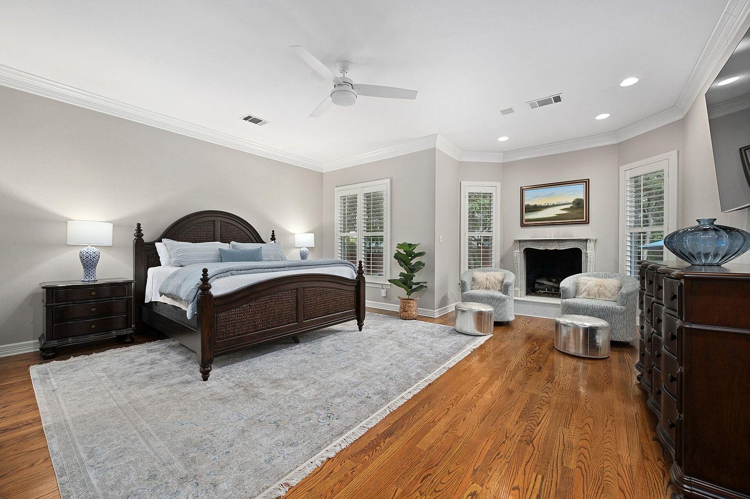 This is a primary bedroom featuring a dark wood, woven-panel bed with light blue accents, a matching nightstand with a blue lamp, and a large area rug. The room also includes a fireplace flanked by windows with plantation shutters, two armchairs with silver ottomans, and a dark wood dresser. The hardwood floors and neutral wall colors create a serene and inviting atmosphere.
