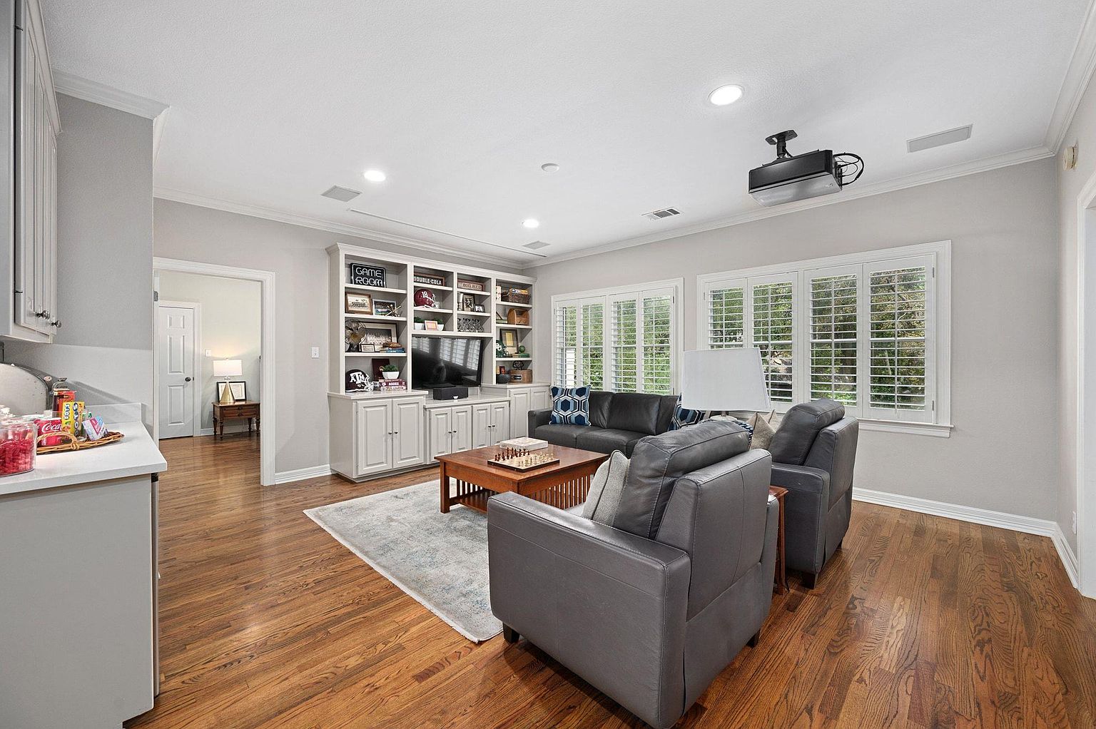 This is an interior shot of a living room featuring hardwood floors, a built-in bookshelf with a television, and two large windows with white shutters. The room is furnished with a dark leather sofa, two gray armchairs, and a wooden coffee table with a chess set. The overall impression is a comfortable and inviting space, perfect for relaxation and entertainment.